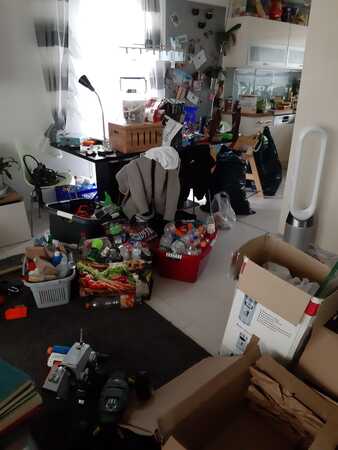 Unordentliche Küche mit vielen Kartons, Haushaltswaren, Spielzeug und vollen Regalen.