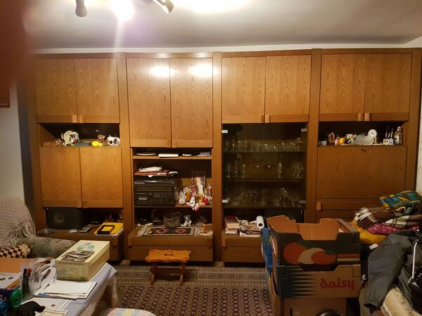 Wohnzimmer mit großer, brauner Schrankwand, Glastüren, Deko, Kartons und Teppich.